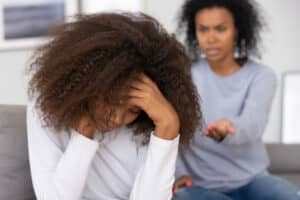 Upset teenage girl rests her head in her hands while her angry mother yells in the background