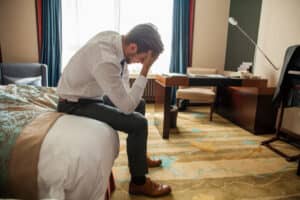 A young businessman in a suit sits on the bed in a hotel room with his face in his palm