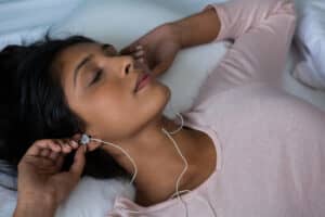 A woman lying down with her eyes closed places earphones in her ears